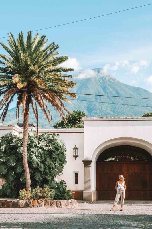 standing infront of a white washed building in antigua guatemala with a palm tree and volcano in the view